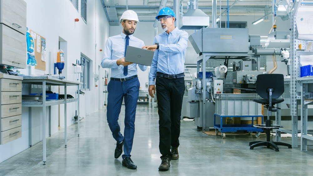 Back View of the Head of the Project Holds Laptop and Discussing Product Details with Chief Engineer while They Walk Through Modern Factory.