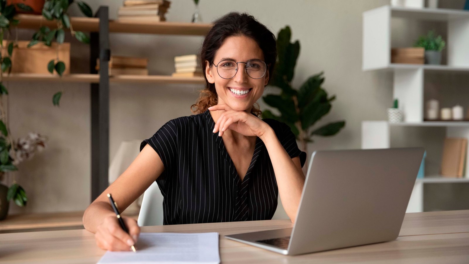 Head shot portrait of smiling woman in glasses writing, taking notes, female student sitting at work desk with laptop, studying online, webinar, confident businesswoman working with documents