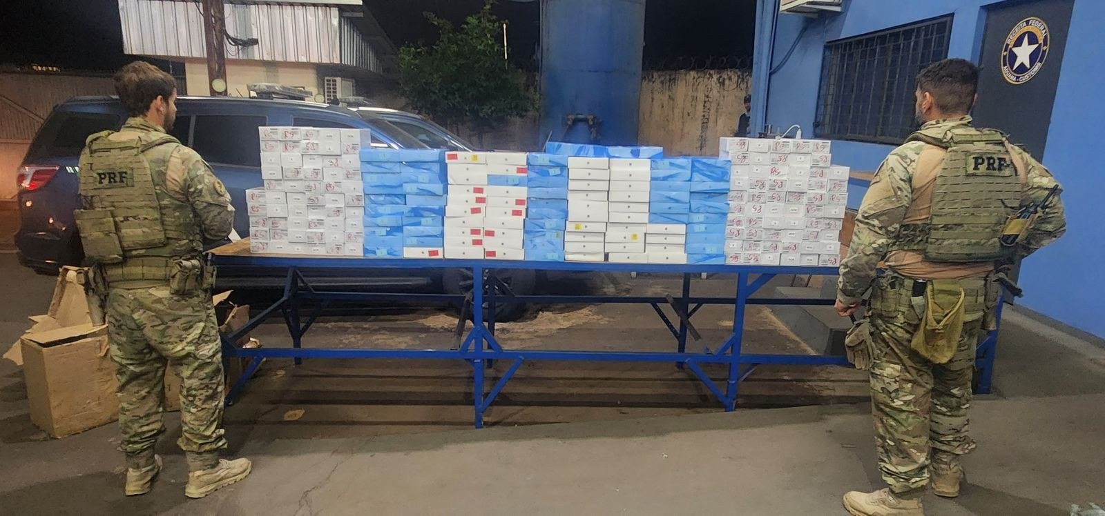 Policiais rodoviários federais ao lado de uma mesa contendo grande quantidade de caixas apreendidas durante ação de fiscalização. Os materiais foram organizados para conferência e registro na unidade policial